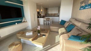 Ein modernes Wohnzimmer mit einem großen Flachbildfernseher, einem beigefarbenen Sofa mit blauen Kissen und einer Küche im Hintergrund.