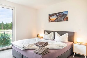 Un dormitorio con una cama grande, una mesita de noche, una lámpara y una ventana que ofrece vistas a árboles y al atardecer.