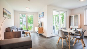 A modern living room with a sofa, dining table, and chairs. There is a wooden cabinet and a TV on a stand. Large windows provide natural light and views of greenery outside. - Apartment Kiefernblick - Haffresidenz (4a.7) - 4a.7 (Zirchow)