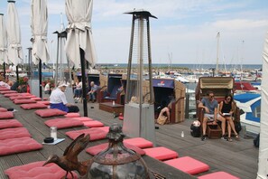Une terrasse dotée de sièges rembourrés rouges, d’une structure métallique imposante et offrant une vue sur les bateaux au loin.