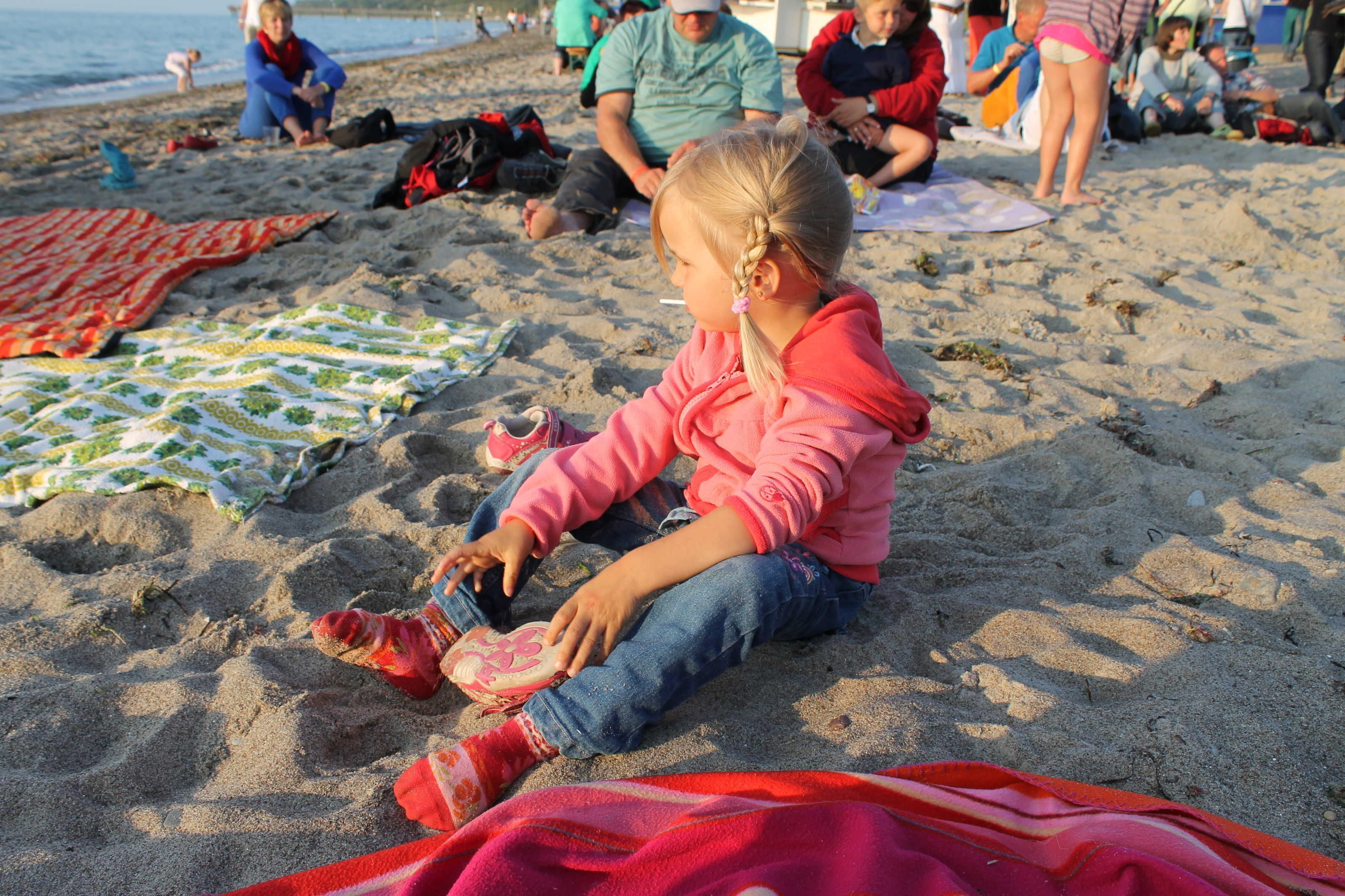 Eine Strandansicht mit Menschen, die auf Decken sitzen, einem Kind, das mit einem Spielzeug spielt, und einem ruhigen Meer im Hintergrund.