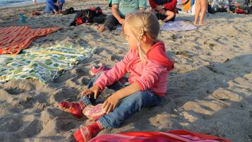 Eine Strandansicht mit Menschen, die auf Decken sitzen, einem Kind, das mit einem Spielzeug spielt, und einem ruhigen Meer im Hintergrund.