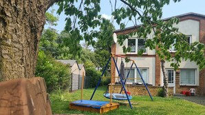 Un banco de madera, un conjunto de columpios y un trampolín en un patio trasero con una casa al fondo.