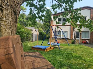 Un banc en bois, une balançoire et un trampoline dans un jardin, avec une maison en arrière-plan.