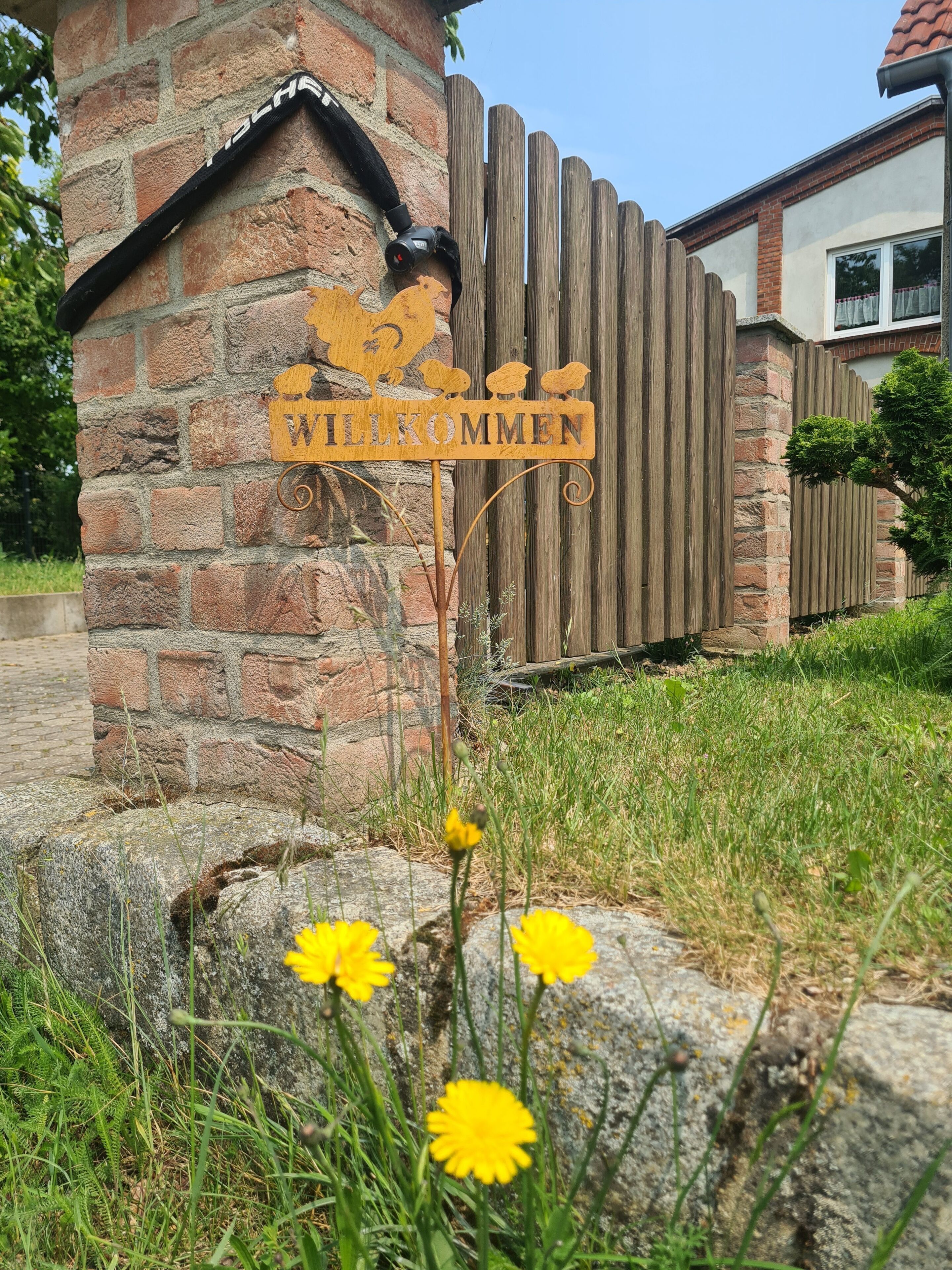 Ein hölzernes Willkommensschild mit Vogelmotiv an einer Ziegelwand.