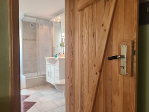 A wooden door with a metal handle, a white bathroom with a sink and mirror, and a shower area.