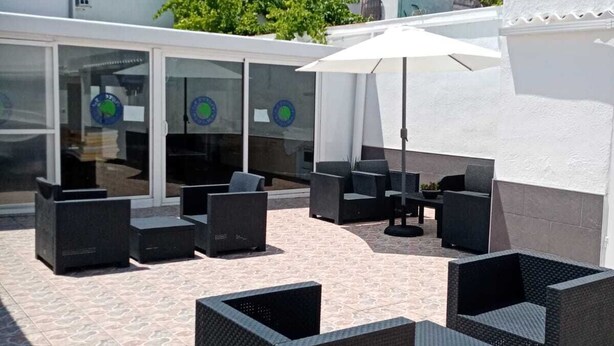 Un espacio de patio al aire libre con mobiliario de mimbre y un paraguas blanco, rodeado por un muro blanco y un ventanal de vidrio con logotipos circulares.
