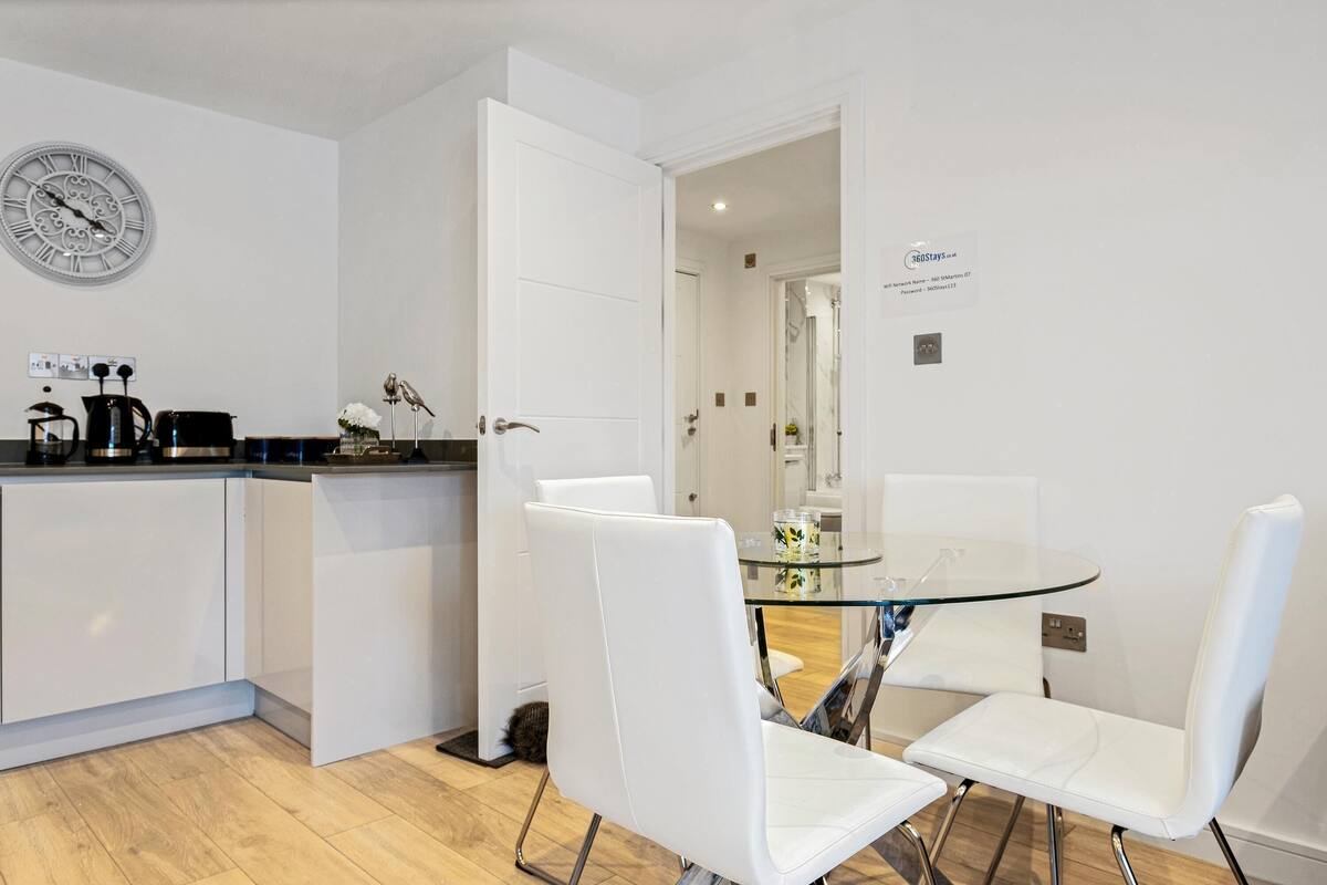 Una cocina moderna con armarios blancos, una cafetera y un reloj en la pared. Hay una mesa de vidrio con sillas blancas en el comedor.