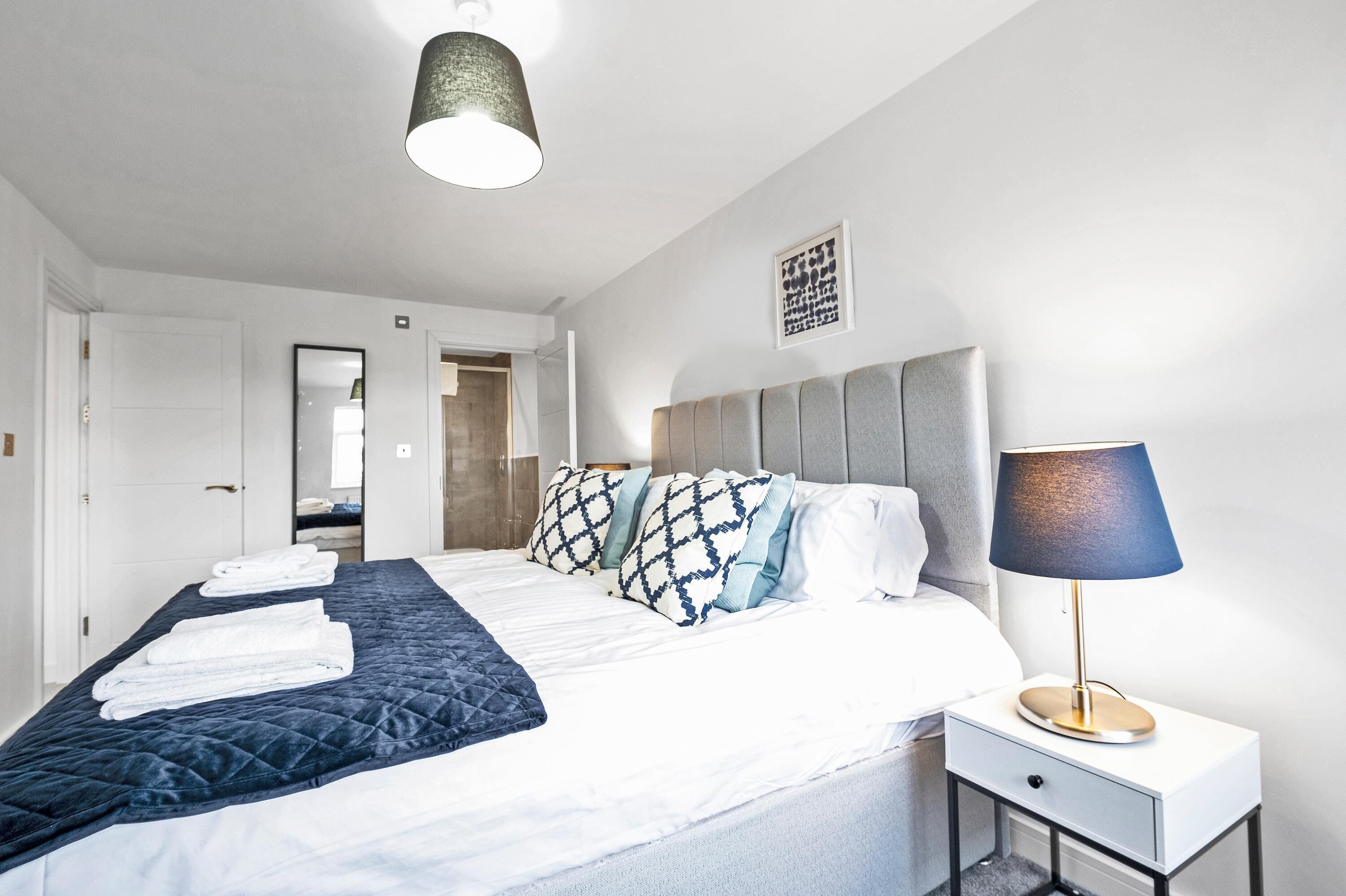 Een opgemaakt bed met een sprei, kussens en een bedlampje. Er is een deur naar een badkamer en een klein tafeltje met een lamp naast het bed.