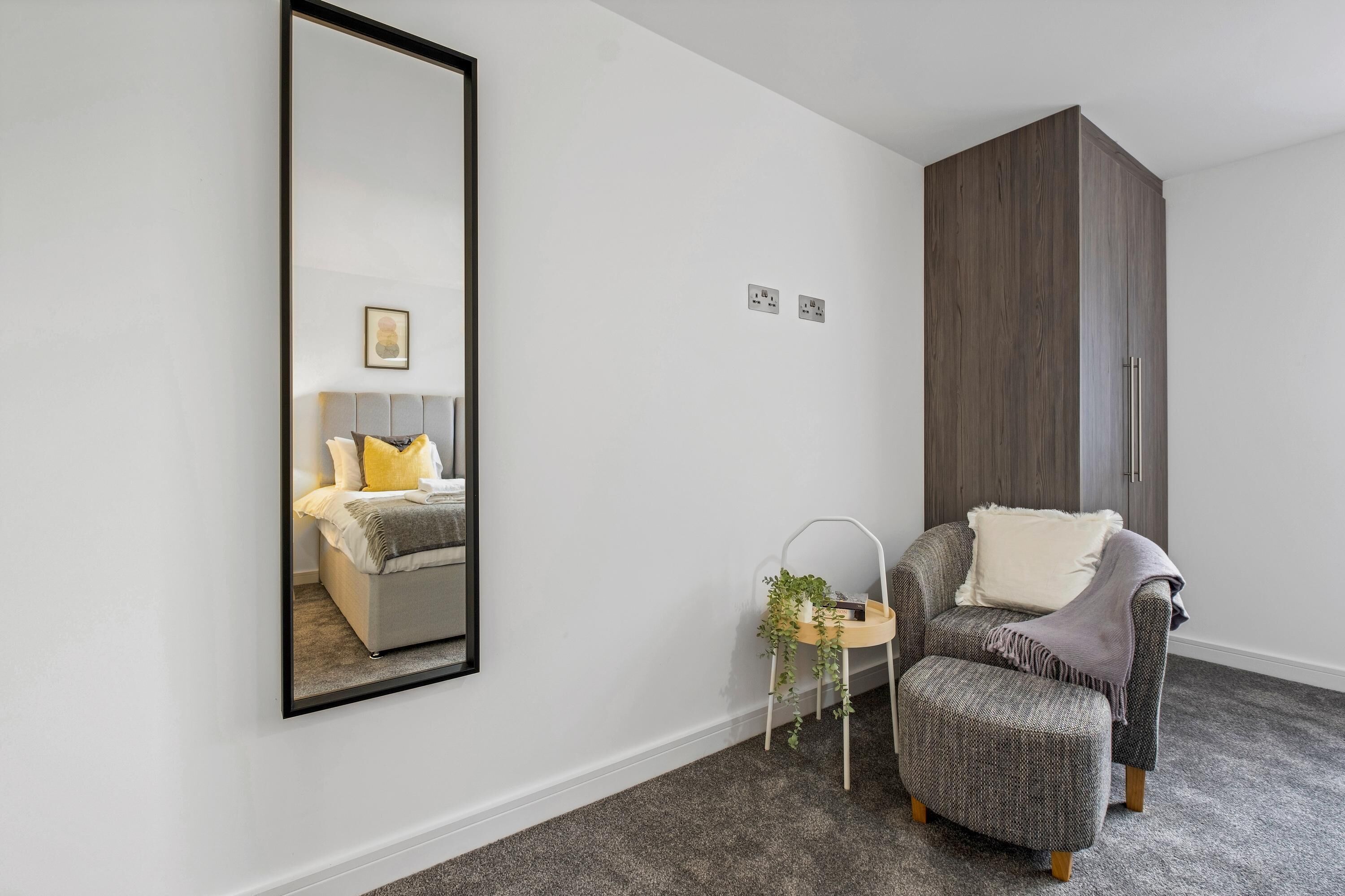 A modern bedroom with a large mirror, a bed with a headboard, a grey upholstered armchair, a small side table with a plant, and a dark wooden wardrobe.