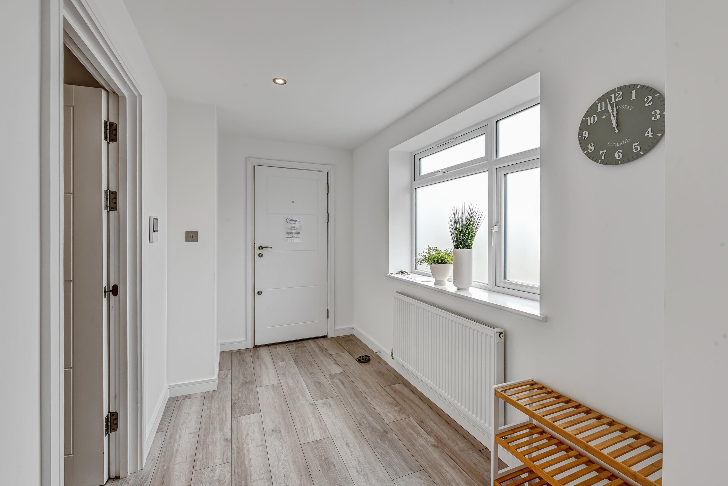 A hallway with a wooden floor, a white door, a window with a view, and a wall-mounted clock.