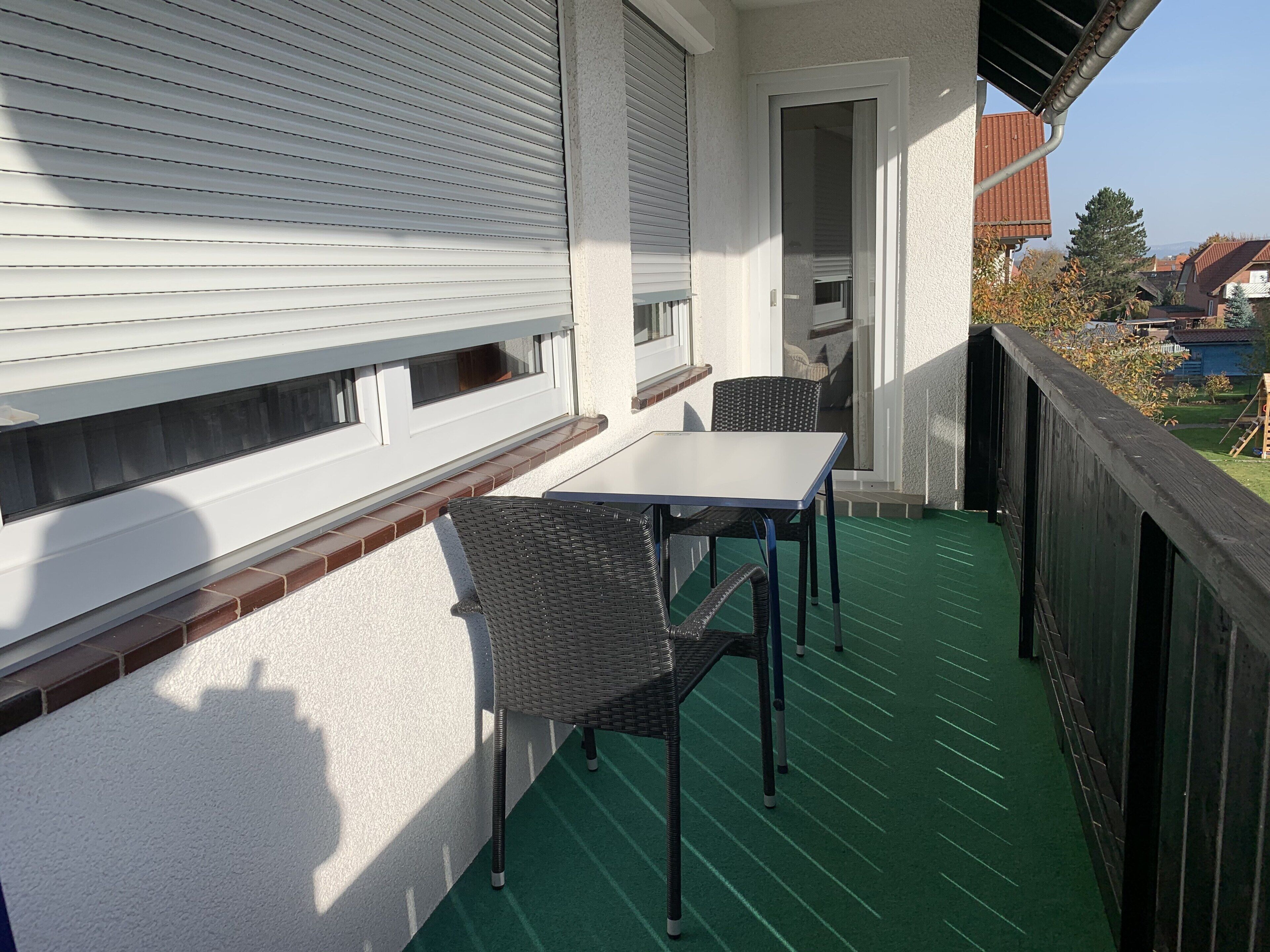 A balcony with a table and two chairs, a sliding door, and a view of the neighborhood.
