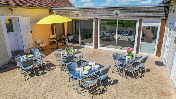 An outdoor patio area with tables and chairs, a yellow umbrella, and a view of a parking lot through large glass doors. - Crazy Villa Luigny (28) - Heated pool - City Stade - 1h20 Paris - 30P (Luigny)