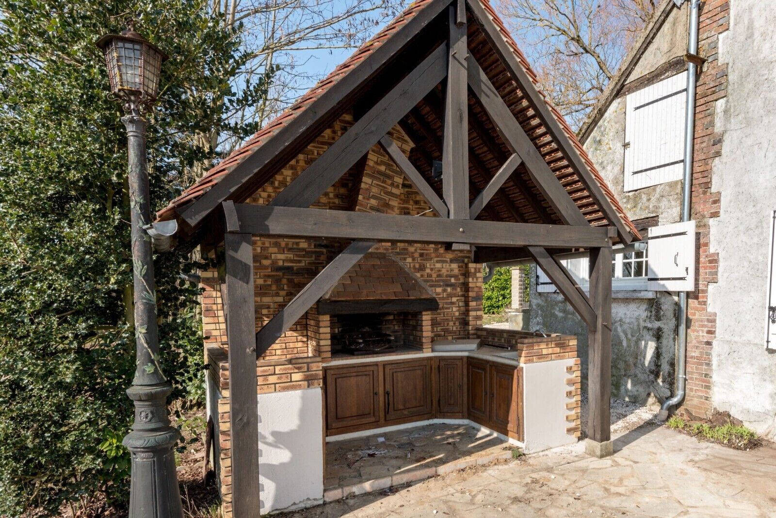 A brick-built outdoor kitchen with a sloped roof, wooden beams, and a stone-paved area.