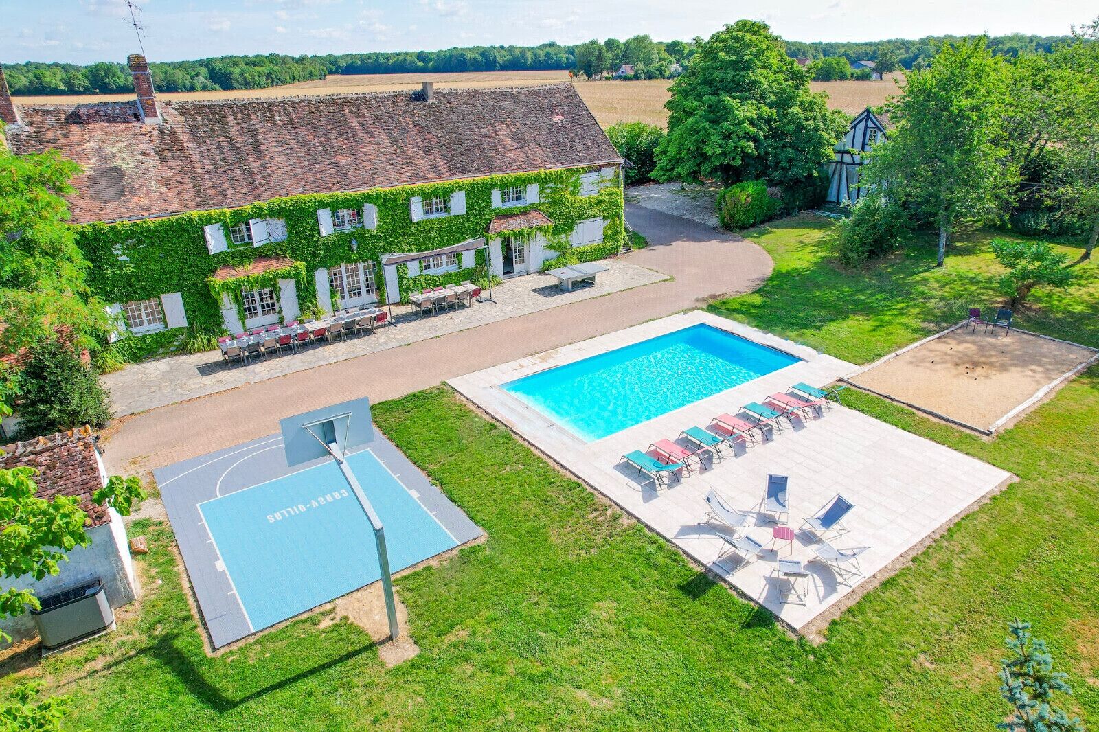 Une grande piscine avec des chaises longues, un terrain de basket et un bâtiment dont les murs sont recouverts de lierre.