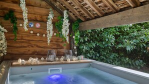 Un jacuzzi con techo y pared de madera. Hay flores blancas colgando del techo y un cartel con el símbolo de prohibido fumar.