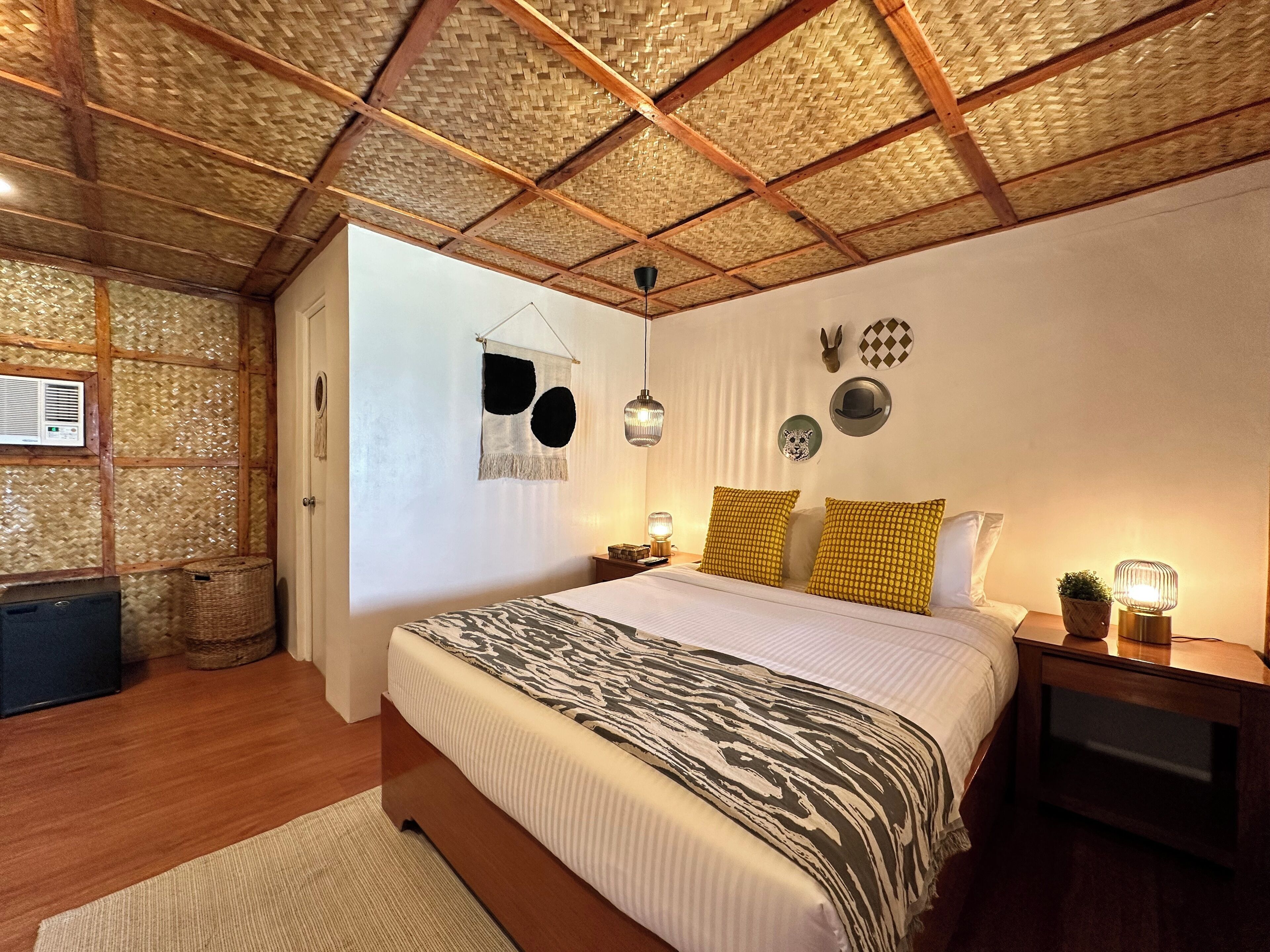 A bedroom with a large bed, wooden beams, a woven ceiling, and a nightstand with a lamp.