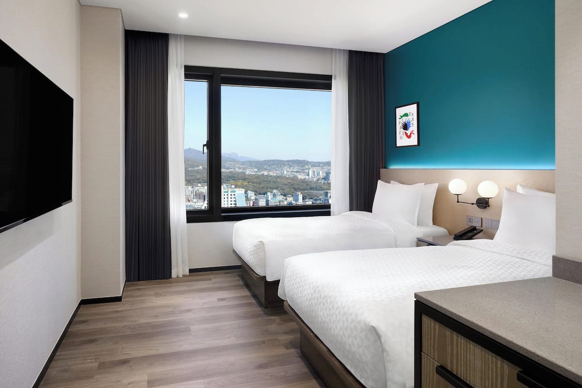 A hotel room with two beds, a large window with a city view, a flat-screen TV, and a small desk with a lamp.
