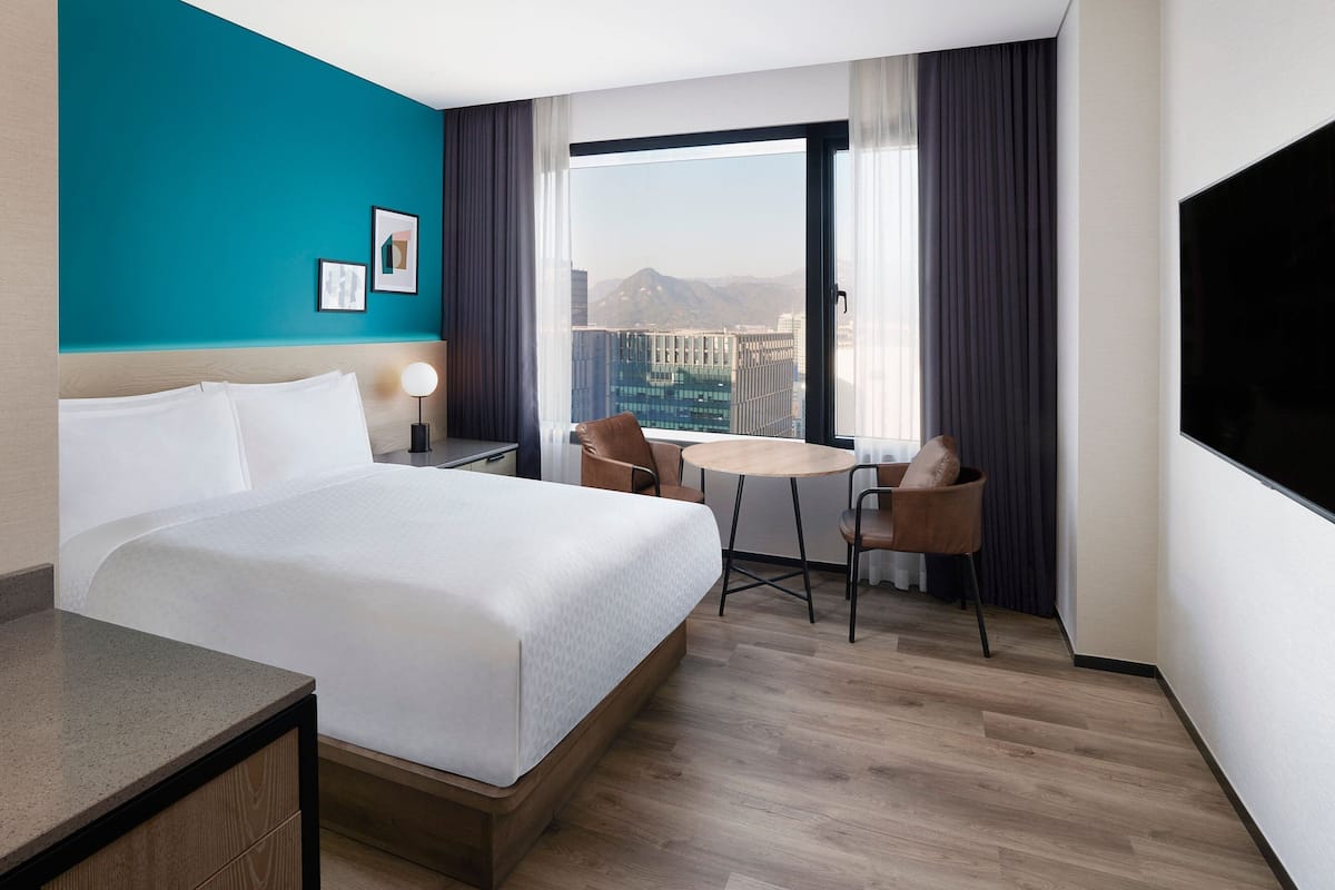 A hotel room with a large bed, a desk, a chair, and a view of a cityscape through a large window.