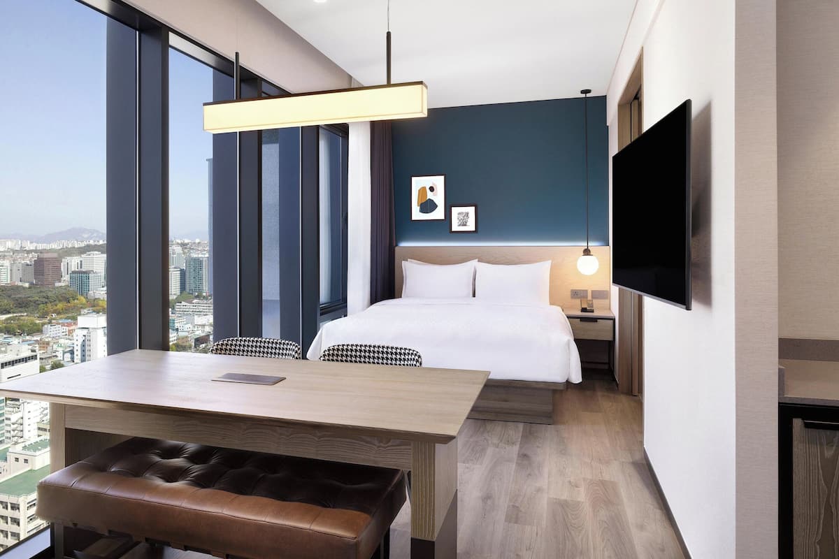 A modern hotel room with a large bed, a desk with a chair, a bench, a TV mounted on the wall, and a city view through large windows.