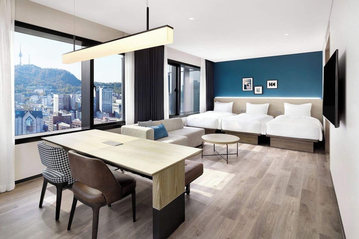 A modern hotel room with a large window offering a city view, a sofa bed, a dining area with a table and chairs, and a small round table.