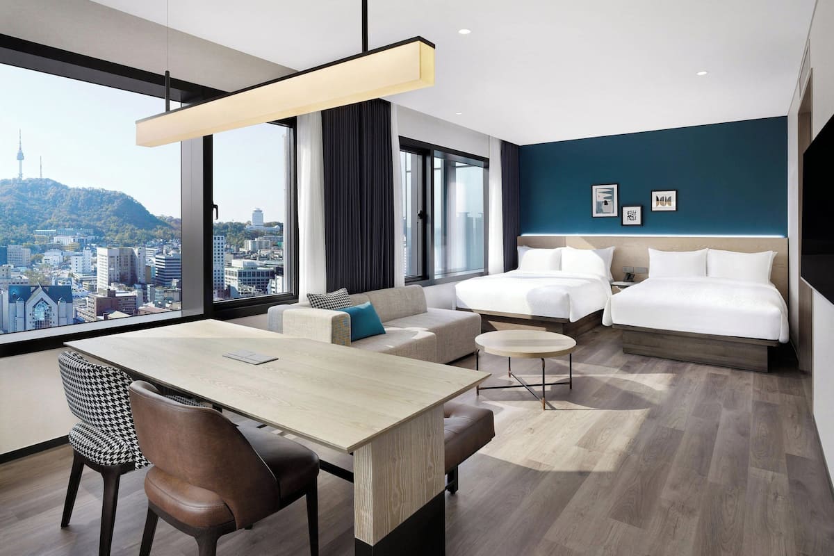 A modern hotel room with a large window, a dining area, and a bed.