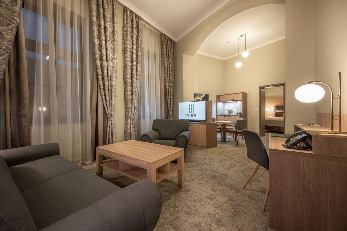 A modern hotel room with a sofa, a wooden coffee table, a TV, a desk with a phone, and a mirror.
