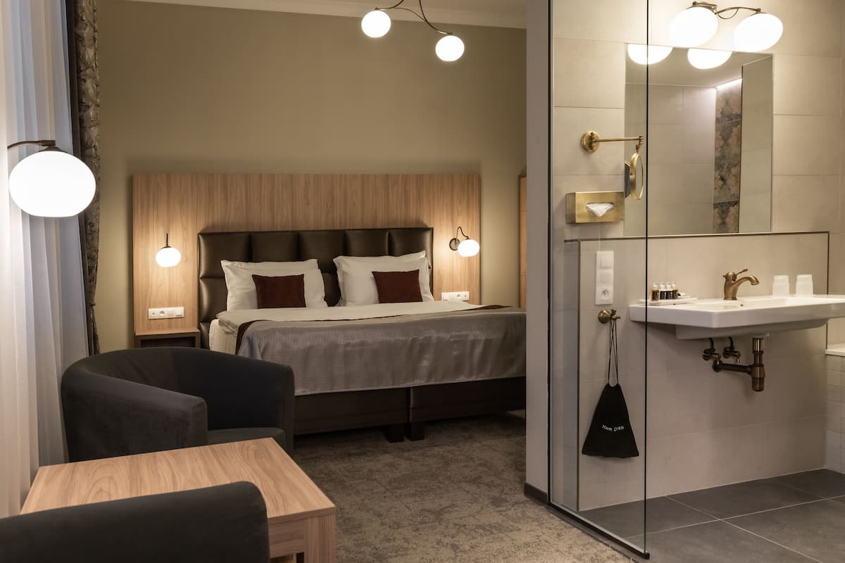 A modern hotel room with a bed, bedside tables, a desk, a chair, a bathroom with a sink and mirror, and a shower.