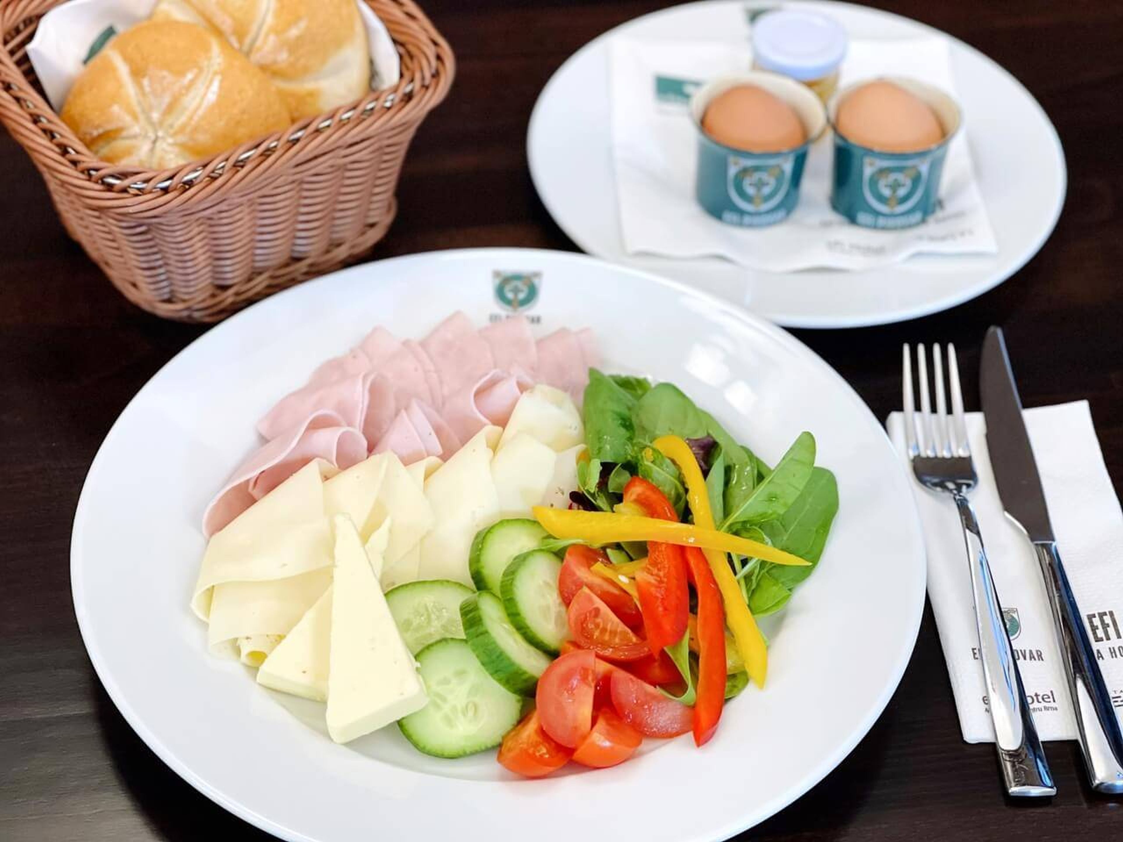 Une assiette avec du jambon, du fromage et une salade de légumes, un panier avec des petits pains, et une assiette avec deux œufs et des condiments.