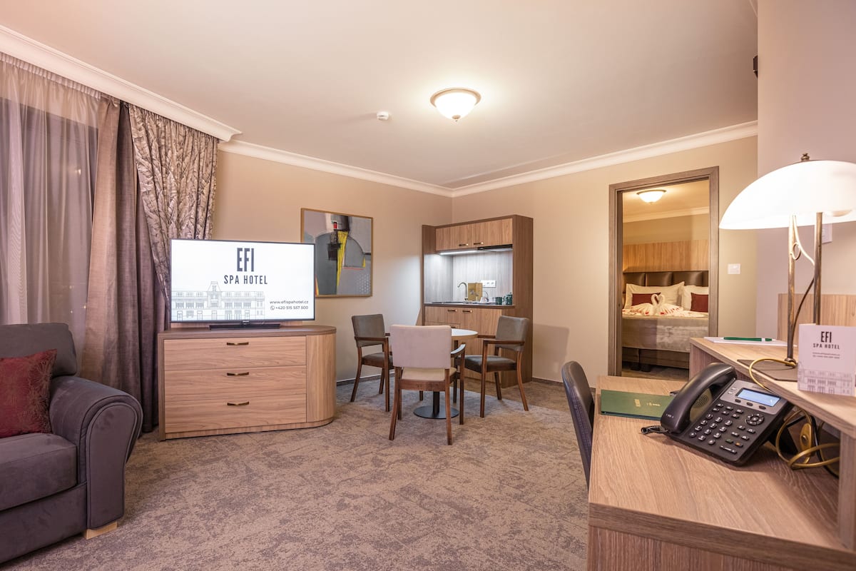 A hotel room with a sofa, a desk with a phone, a TV, a dining area with chairs, a kitchen, and a bathroom.