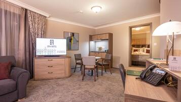 Habitación de hotel con sofá, escritorio con teléfono, televisor, zona de comedor con sillas, cocina y baño.