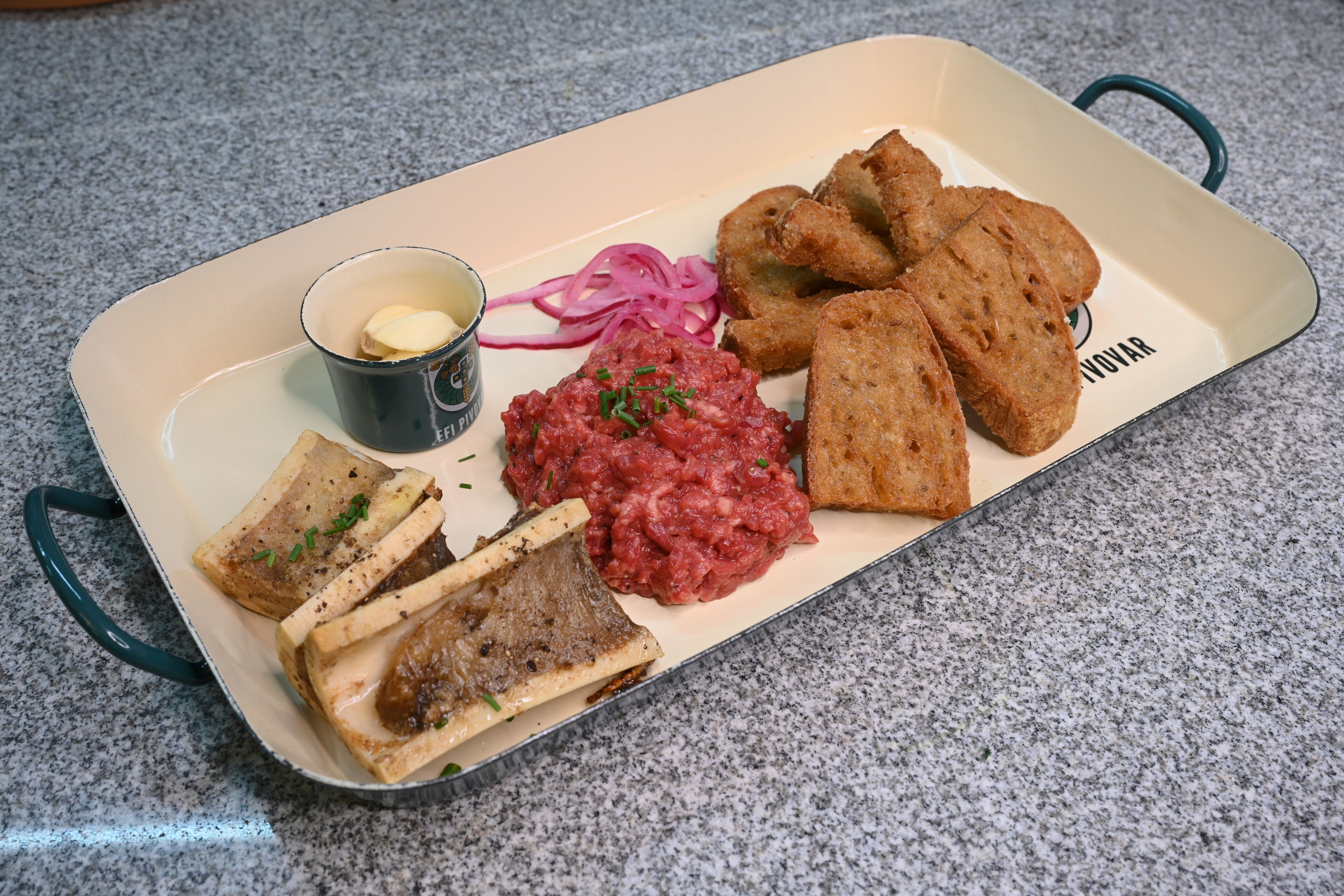 A rectangular serving tray with a variety of food items including bread, a red sauce, pickled onions, and a small cup of butter.