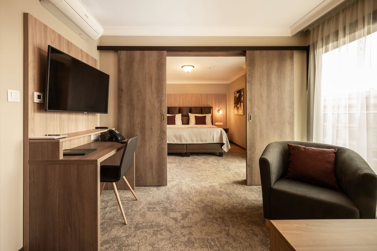 A hotel room with a wooden desk, a flat-screen TV, a bed with a beige bedspread, a dark-colored armchair, and a large window with curtains.