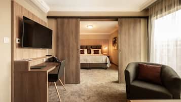 A hotel room with a wooden desk, a flat-screen TV, a bed with a beige bedspread, a dark-colored armchair, and a large window with curtains.
