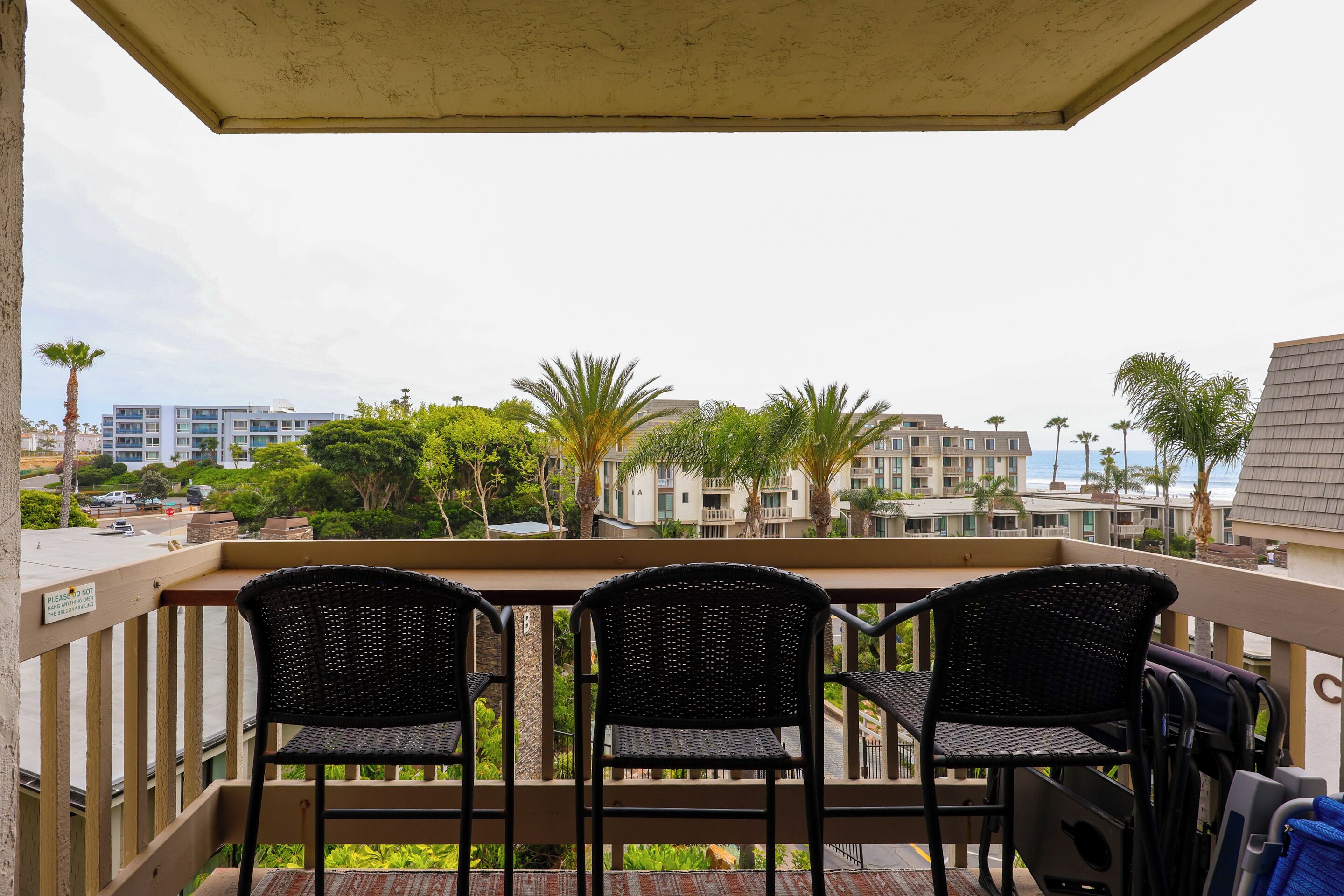 A balcony with three wicker chairs, a grill, and a view of buildings and palm trees.