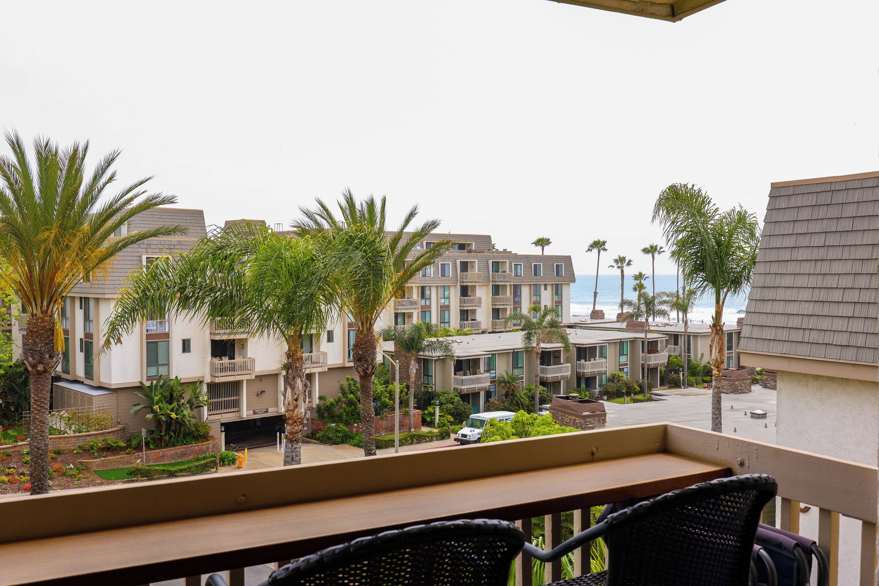 A balcony with wicker chairs, a view of a hotel building with multiple balconies, and a parking area with a white vehicle.