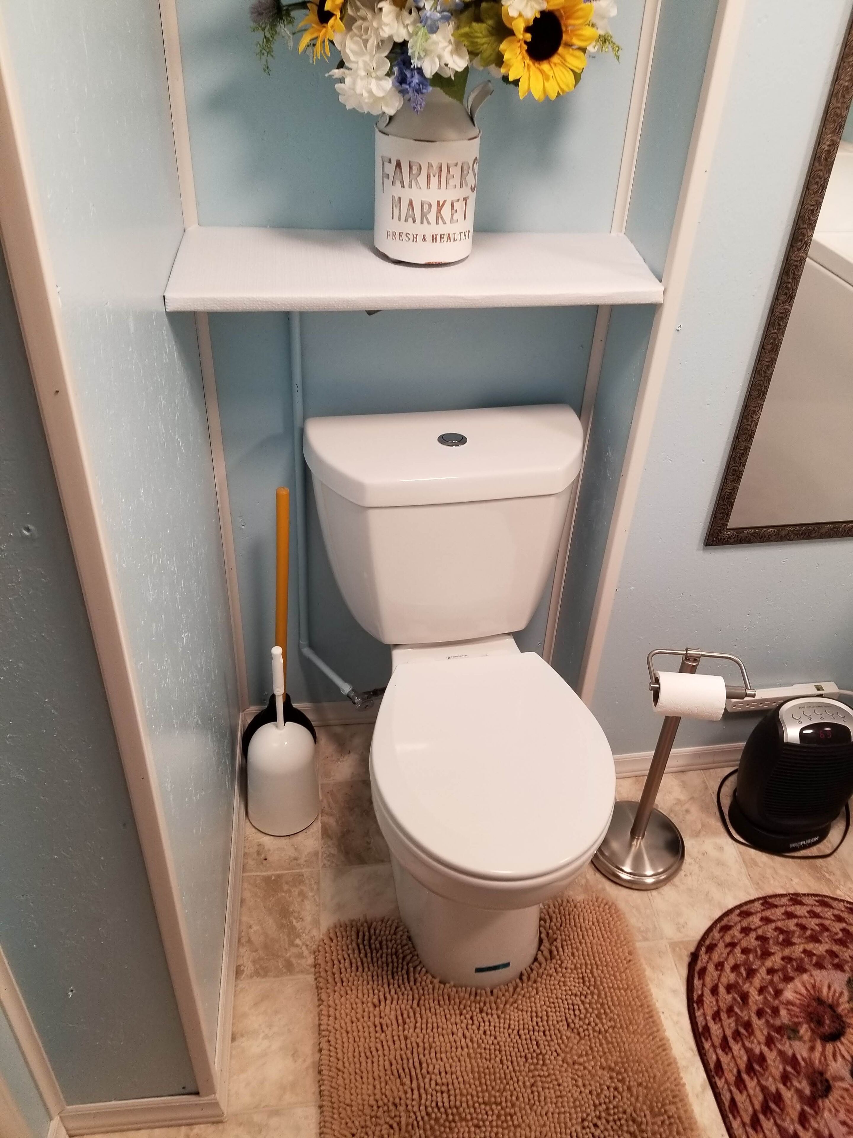 A bathroom with a toilet, a toilet brush, a paper towel holder, and a floor mat.