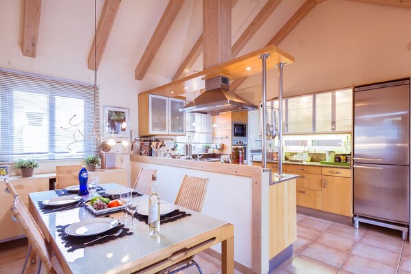 Una cocina moderna con vigas de madera, una isla central, electrodomésticos de acero inoxidable y un comedor con una mesa para dos.