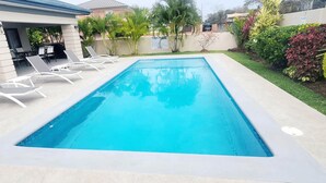Una piscina rectangular con agua azul cristalina, rodeada por una terraza de concreto. Hay sillas de descanso blancas alineadas a lo largo del borde de la piscina.