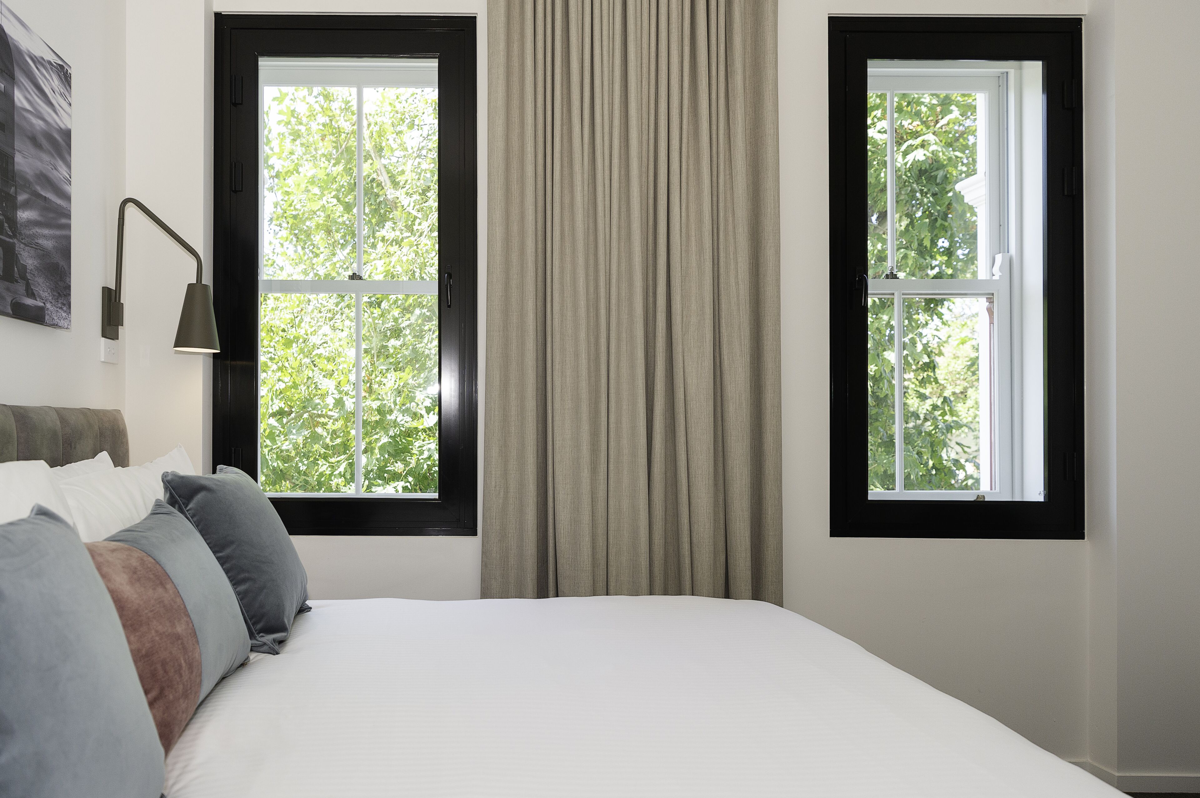 A bedroom with a bed, two windows, a lamp, and a painting on the wall.
