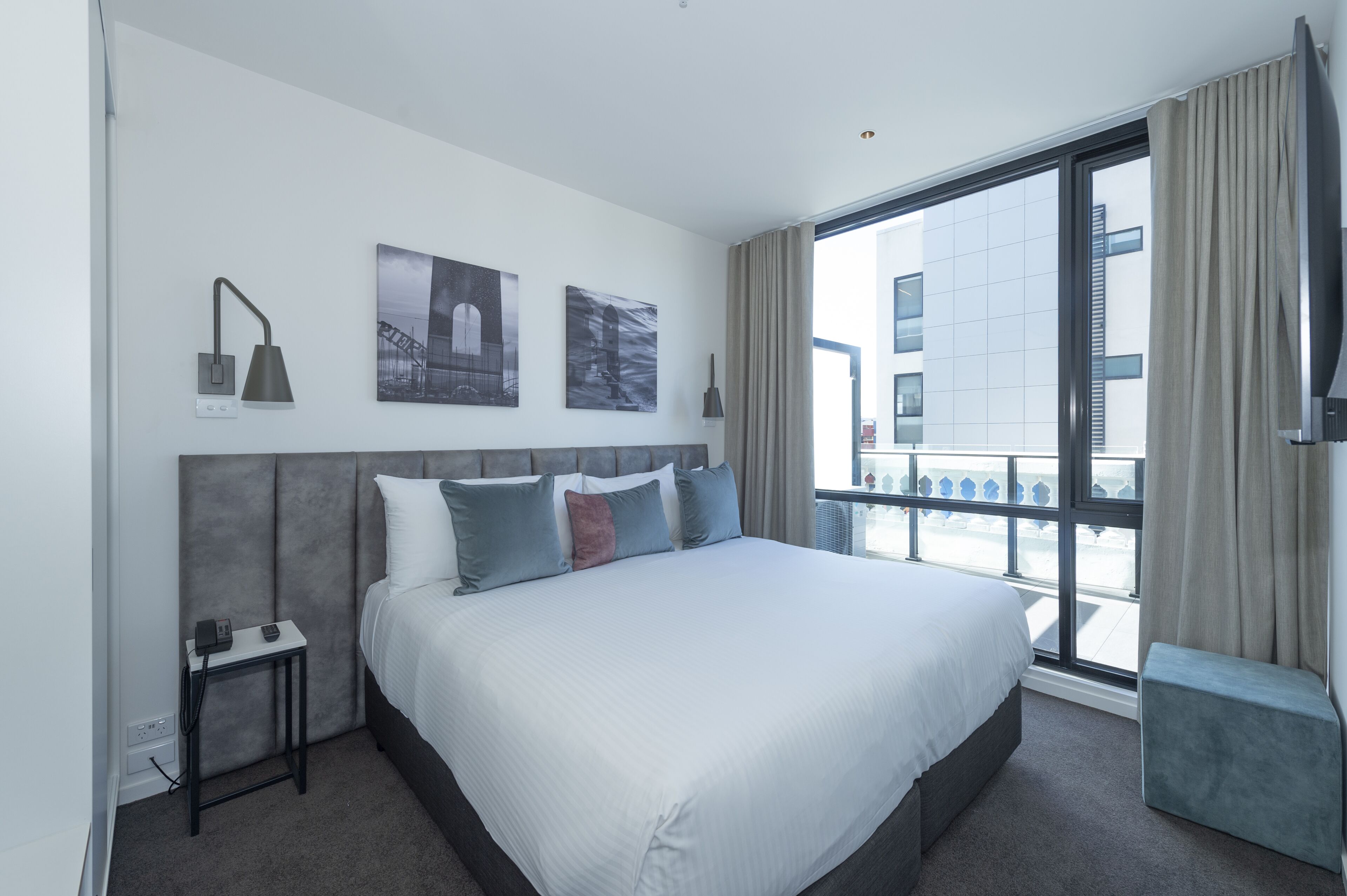 A modern hotel room with a large bed, a side table, a lamp, and a window with a view of buildings.