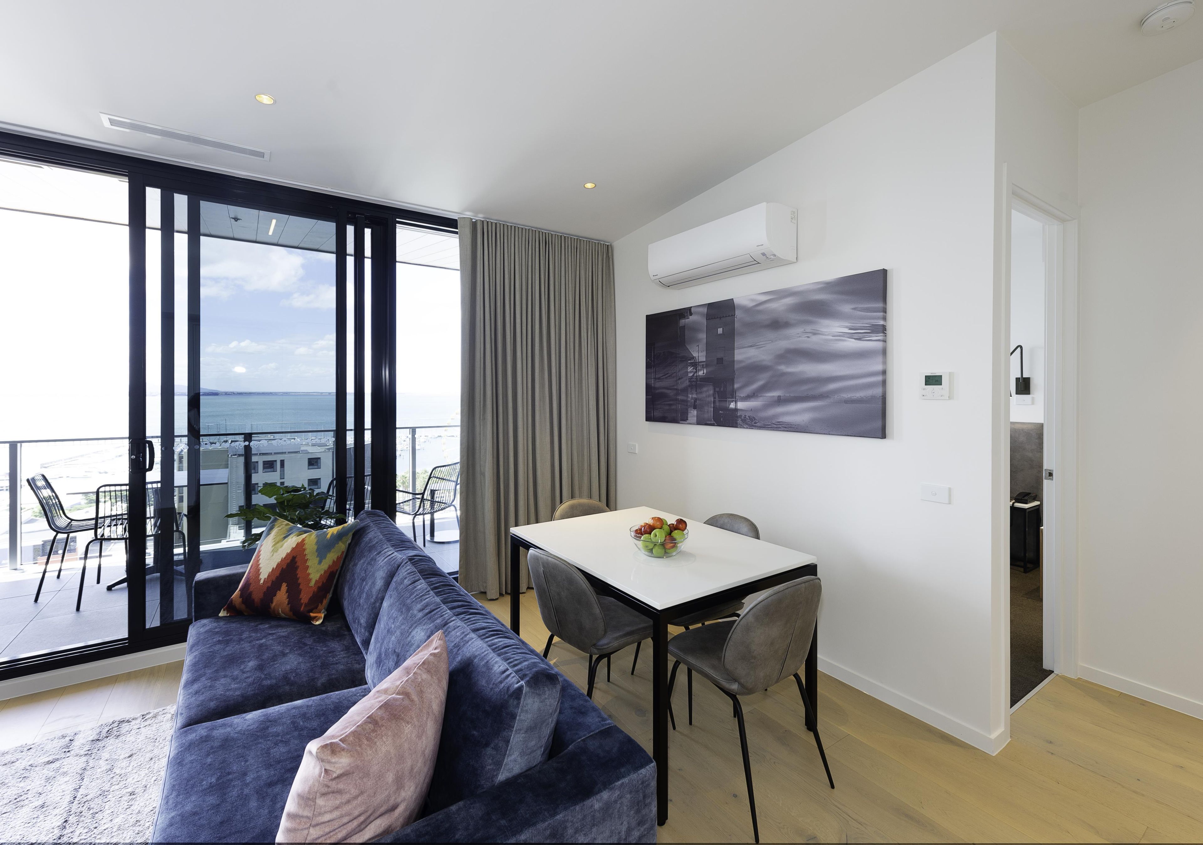 A modern living room with a sofa, a dining table with chairs, a view of the sea, and a mounted air conditioner.