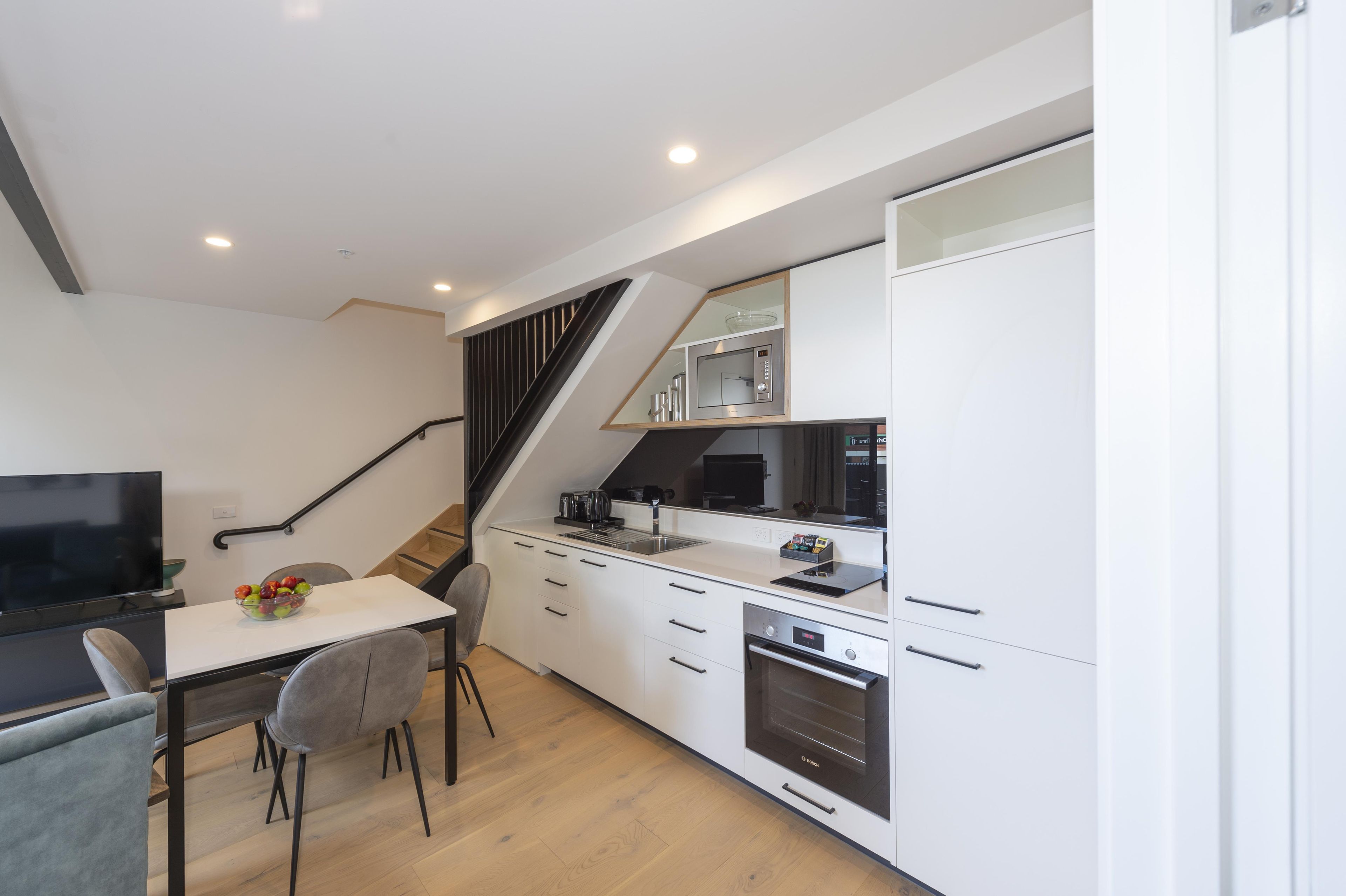 A modern kitchen with a microwave, oven, and a dining area with a TV and a bowl of fruit.