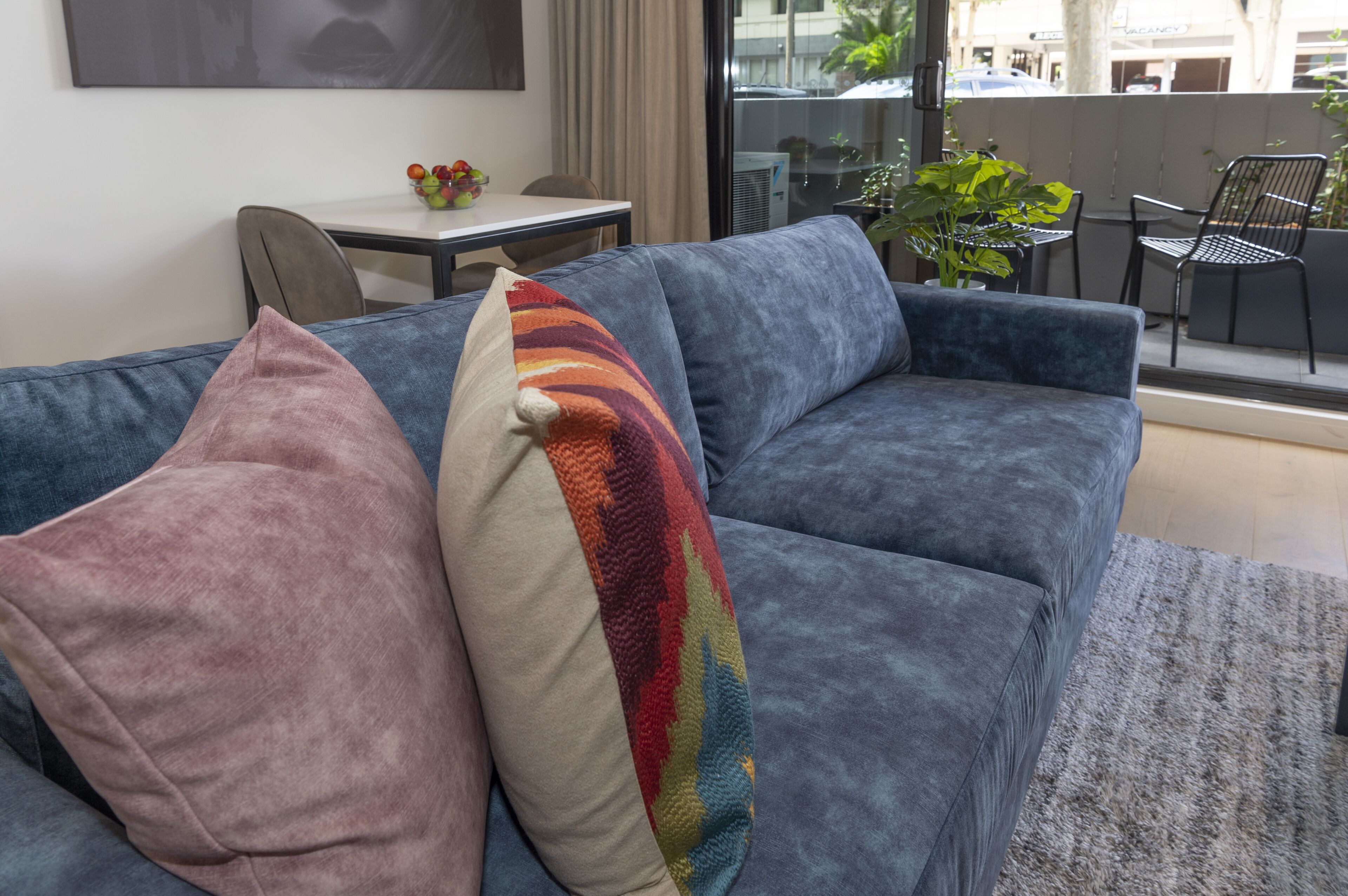 A modern living room with a grey sofa, two decorative pillows, a small table with fruit, and a view of the outdoors.