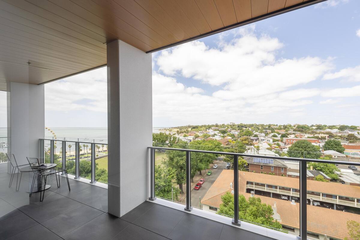 A balcony with a view of a residential area, a table with chairs, and a clear sky.