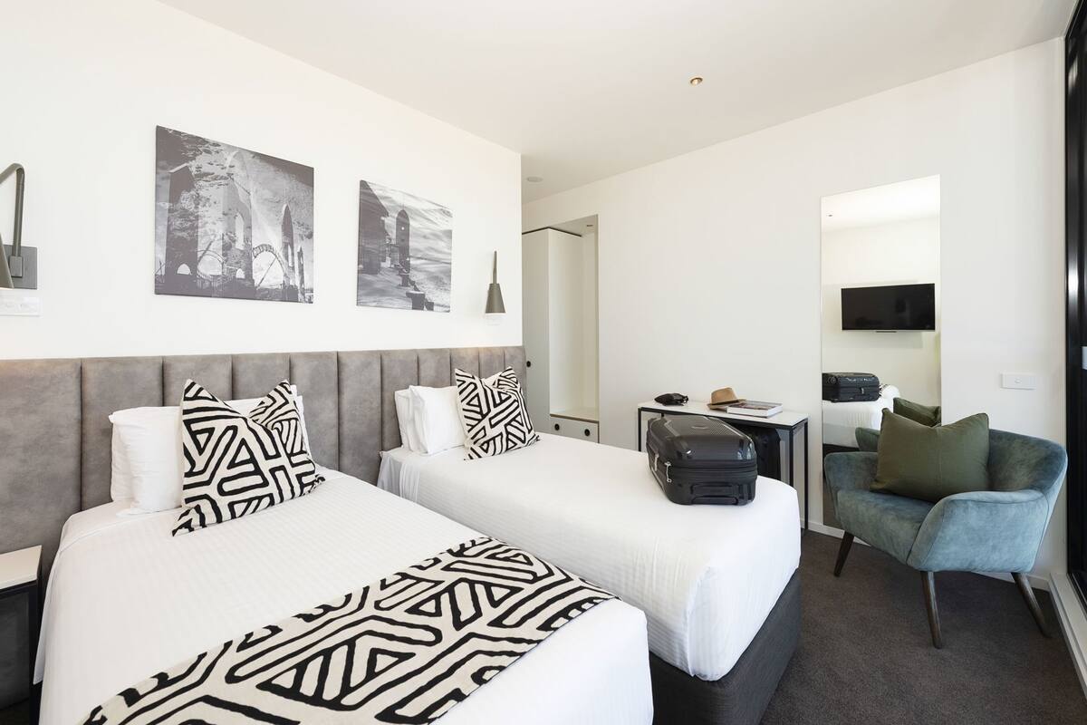 A hotel room with two beds, a desk, a chair, a TV, and artwork on the wall.