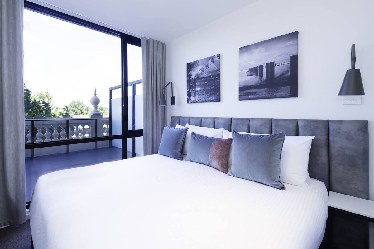 A modern hotel room with a large bed, two wall-mounted artworks, a balcony with a railing, and a large window.