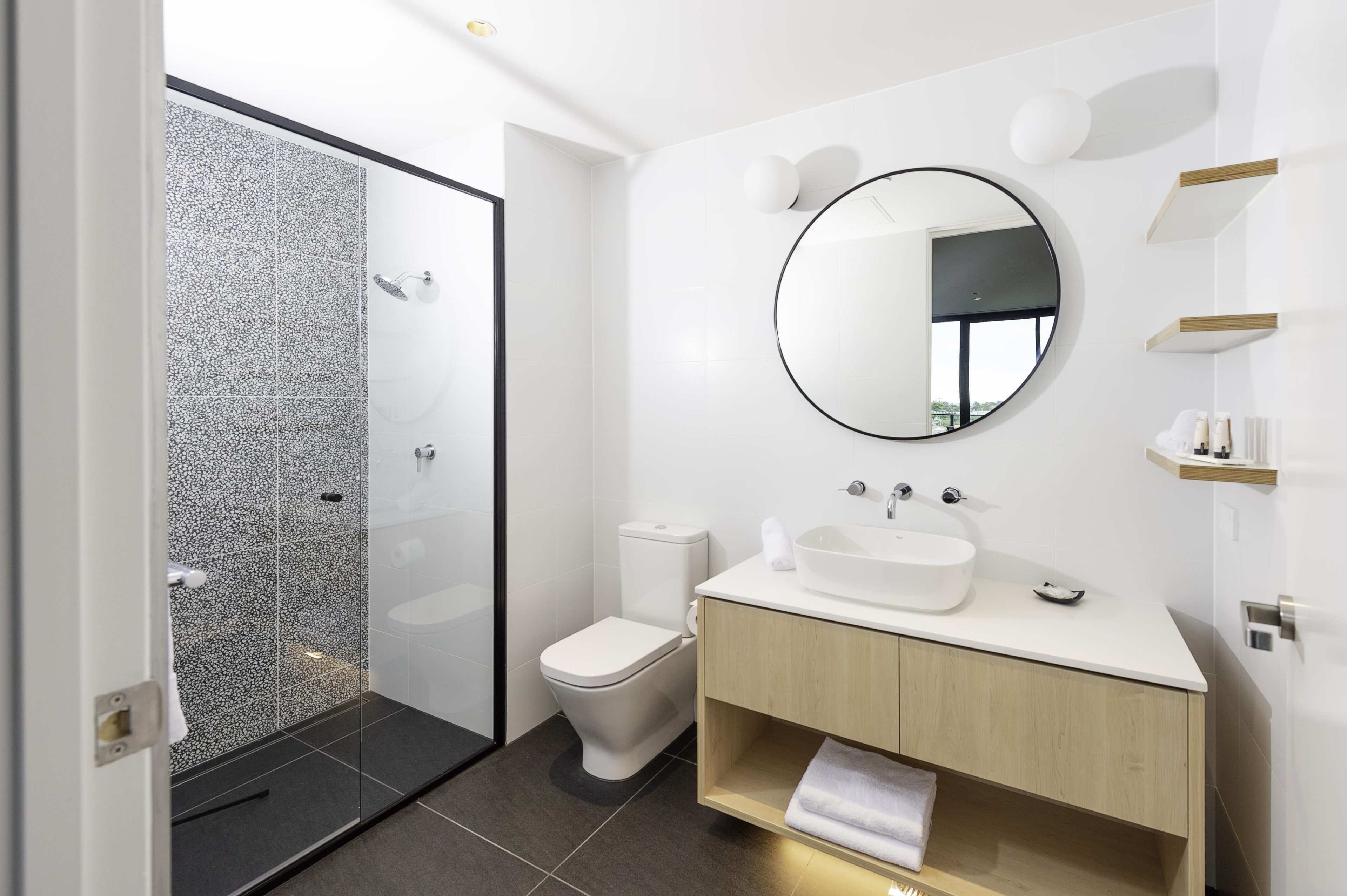A modern bathroom with a glass shower enclosure, a white toilet, and a vanity with a round mirror.