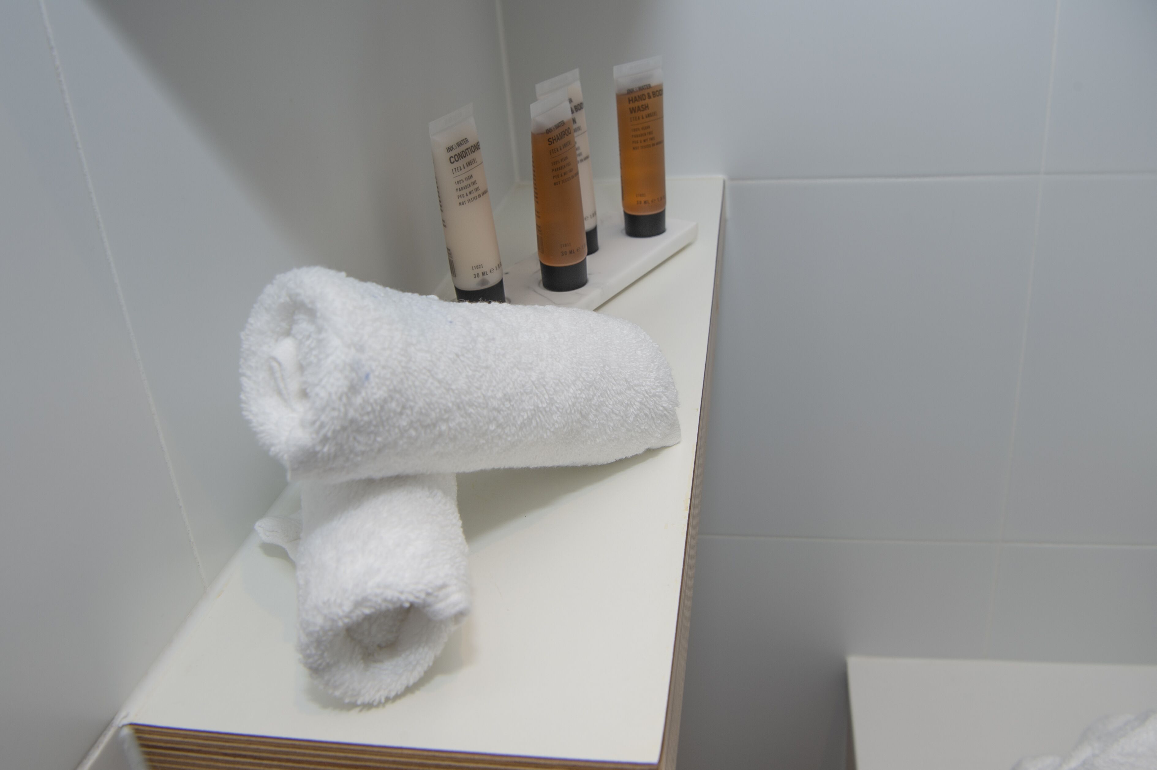 Ett badrum med en vit handduk på en hylla, tre flaskor med hygienprodukter och en vit kakelbeklädd vägg.