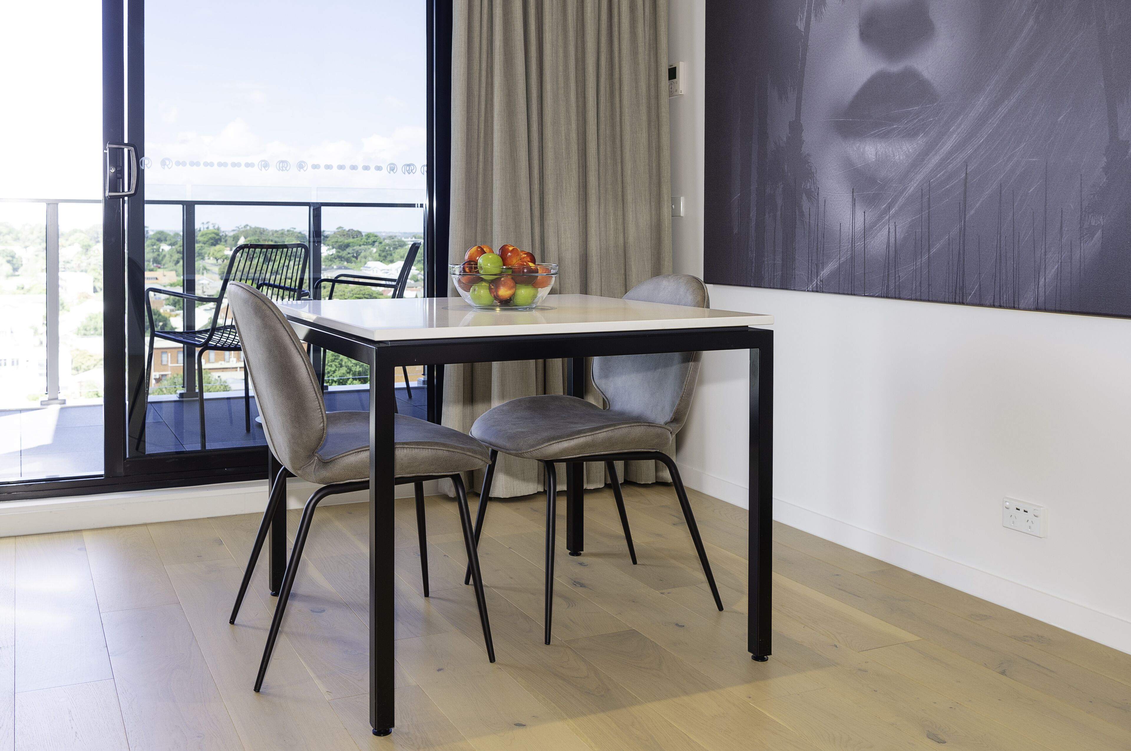 A dining area with a table and chairs, a bowl of fruit, and a view of greenery through large windows.